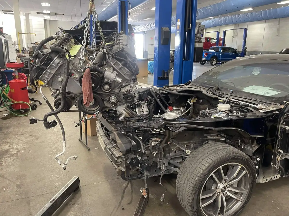 Coyote V8 engine suspended from a hoist beside a stripped Mustang engine bay